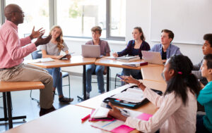 High School Teacher Talking To Pupils Using Digital Devices In Technology Class